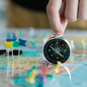 Close-up of hand holding compass over a map with colorful pins and flags, symbolizing travel planning.