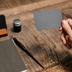 A close-up of a hand holding a blank business card above a wooden desk with office accessories.